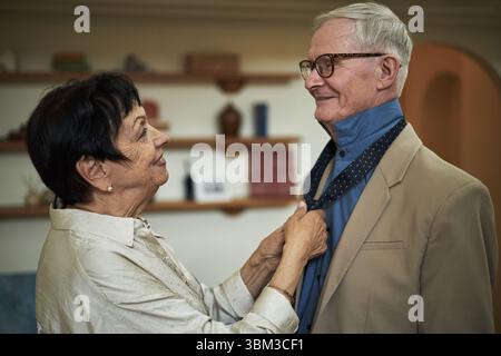 Älteres Paar genießt einen zärtlichen Moment, als Frau einem Mann mit Krawatte hilft und ein echtes Lächeln teilt. Im Hintergrund gibt es ein Bücherregal mit verschiedenen Gegenständen, die eine gemütliche Atmosphäre schaffen Stockfoto
