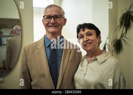 Porträt eines älteren Ehepaares, das im Haus lächelt und mit entspanntem Ausdruck steht, elegant für Partys oder Konzerte gekleidet. Ein Gentleman trägt einen Anzug, während die Dame in der Bluse ist Stockfoto