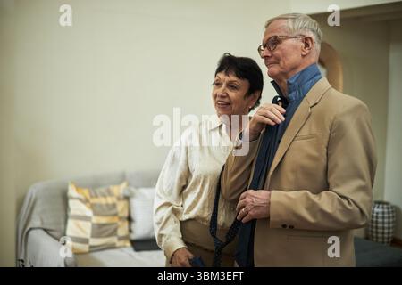 Älteres Ehepaar, das in einem gemütlichen Wohnzimmer steht und zufrieden und komfortabel aussieht. Beide tragen legere Outfits und Brillen mit weichen Möbeln im Hintergrund Stockfoto