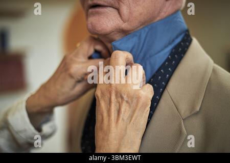 Nahaufnahme von weiblichen Händen, die den blauen Hemdkragen für ältere Herren mit Anzug und Befestigungskrawatte anpassen. Das Gesicht eines älteren Mannes ist teilweise sichtbar und zeigt Sorgfalt und Aufmerksamkeit Stockfoto