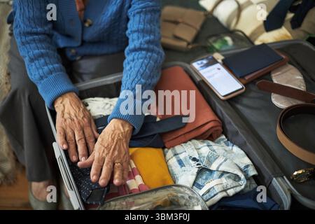 Senior Person packt Gepäck mit verschiedenen wichtigen Dingen, einschließlich Kleidung, Gadgets und Dokumenten, und bereitet sich auf die Reise im Urlaub vor. Konzentrieren Sie sich auf die Hände, um Gegenstände im Koffer mit Sorgfalt zu organisieren Stockfoto