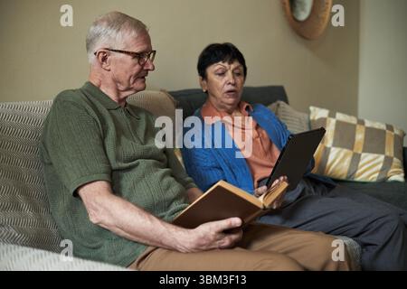 Seniorenpaar, das auf dem Sofa sitzt und mit einem Tablet und einem anderen im gemütlichen Wohnzimmer mit einem Buch liest. Spiegel und gemusterte Kissen zieren den Hintergrund Stockfoto