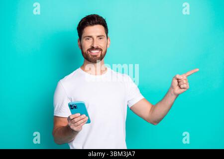 Glücklicher Mann, der ein Smartphone hält, während er auf etwas aufregendes zeigt und ein Konzept oder einen Service vor blauem Hintergrund fördert Stockfoto