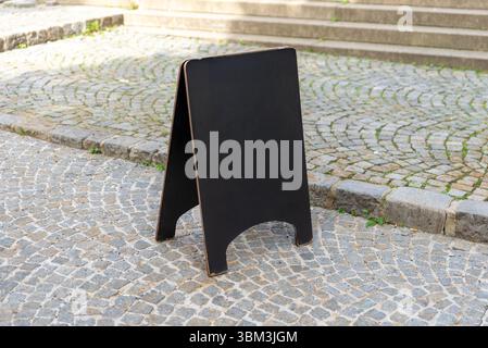 Schwarzes Schild mit Tafel auf der Kopfsteinpflasterstraße, ideal für Cafés, Restaurants und lokale Angebote in urbanen Outdoor-Umgebungen Stockfoto