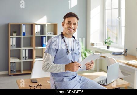 Porträt eines jungen glücklichen männlichen Arztes in blauer medizinischer Uniform, der am Schreibtisch in seinem Büro sitzt. Stockfoto