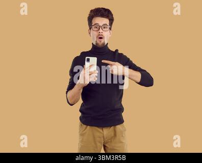 Der staunende junge Brünette mit schwarzem Rollkragenkragen zeigt auf beigefarbenem Hintergrund auf das Smartphone. Stockfoto