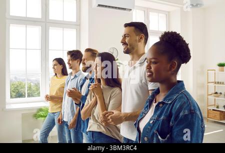 Fröhliche junge, abwechslungsreiche Gruppe in legeren Kleidern, die drinnen in einer Reihe stehen und lächeln. Stockfoto