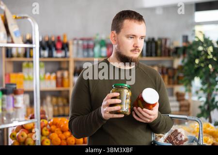 Der Mann wählt Tomaten und Gurken aus der Dose im Supermarkt sorgfältig aus Stockfoto