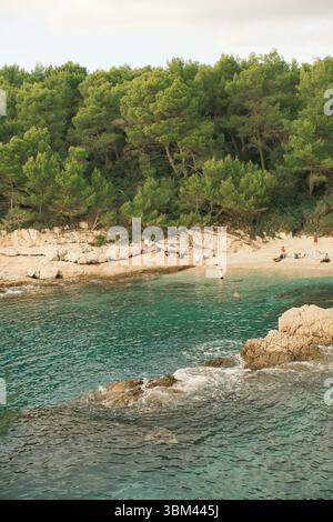 Idyllische Bucht mit Menschen, die im türkisfarbenen Wasser schwimmen, umgeben von felsigem Ufer und dichtem Kiefernwald. Friedliche mediterrane Landschaft. Stockfoto