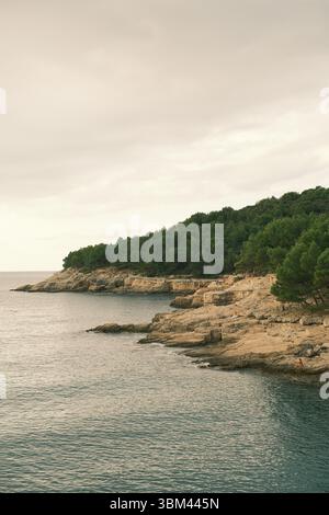 Dramatische Adriaküste mit bewaldeten Klippen und felsigem Strand bei Sonnenuntergang. Touristen, die sich sonnen und die natürliche Küste erkunden. Stockfoto