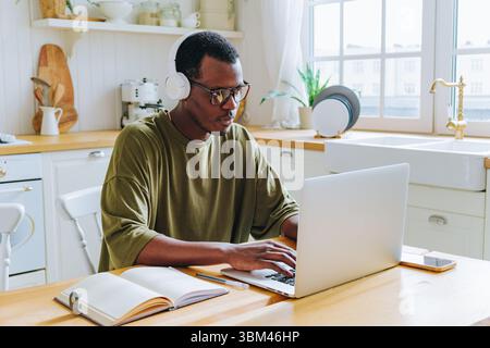 Der junge afroamerikanische Programmierer, der Kopfhörer und Brillen trägt, konzentriert sich auf seine Arbeit an einem Laptop in einer hellen Küche und demonstriert so den modernen Lebensstil der Arbeit von zu Hause aus Stockfoto