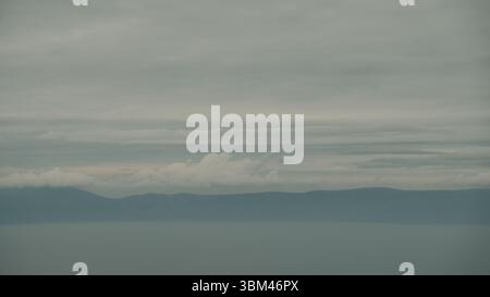 Das ruhige Wasser der Adria verschmilzt nahtlos mit den nebeligen Silhouetten der fernen Berge, die unter schweren, mehrschichtigen Wolken verborgen sind. Stockfoto