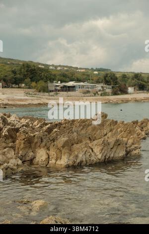 Kalksteinformationen an der Küste führen das Auge in Richtung einer kleinen adriatischen Stadt mit niedrigen Häusern, die zwischen Bäumen eingebettet sind. Ruhiges Meer, bedeckter Himmel. Stockfoto
