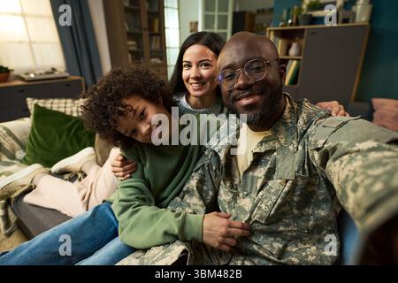 Porträt eines schwarzen Mannes mittleren Alters in Militäruniform, der auf dem Sofa sitzt, mit lächelnder kaukasischer Frau und einem lockigen Kind Stockfoto