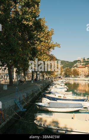 Eine ruhige Stadtpromenade erstreckt sich entlang eines ruhigen Kanals, an dem kleine Boote ankern und sanft in der Mittagssonne schaukeln. Stockfoto