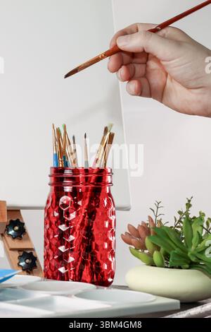 Hand des Künstlers, der den Pinsel neben Mauerglas und Leinwand hält – Creative Workspace Scene Stockfoto