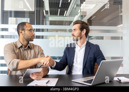 Zwei Geschäftsleute schütteln sich in einem modernen Büroumfeld die Hand, wahrscheinlich nach einem erfolgreichen Abschluss oder einer erfolgreichen Vereinbarung. Konzentrieren Sie sich auf Zusammenarbeit und Teamarbeit. Stockfoto