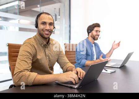Zwei verschiedene Kundendienstmitarbeiter, die Headsets tragen, arbeiten in einer modernen Büroumgebung an Laptops. Stockfoto