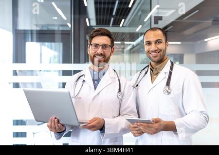 Zwei lächelnde Ärzte posieren in einem modernen Büro, einer hält einen Laptop und der andere ein Tablet. Stockfoto