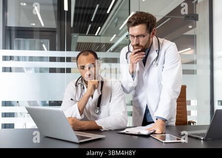Zwei Ärzte, die weiße Mäntel und Stethoskope tragen, betrachten einen Laptop-Bildschirm in einer modernen Büroumgebung, tief in Gedanken. Stockfoto