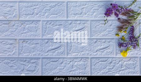 Helle Komposition von Wildblumen, einschließlich gelber und lila Blüten, in der unteren rechten Ecke einer weißen strukturierten Ziegelwand. Minimalisti Stockfoto