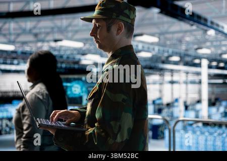 Soldat, der sichere militärische Netzwerke innerhalb des Rechenzentrums pflegt. Militär-IT-Experte in der Überwachung von geheimen Systemen im Serverraum mithilfe eines Laptops Stockfoto
