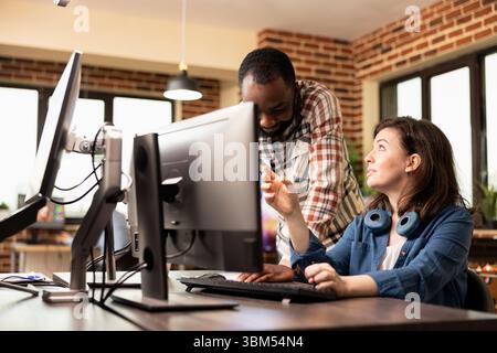 Schwarzer männlicher Manager, der am Schreibtisch steht und Produktdesigns mit einem kaukasischen Mitarbeiter diskutiert, der in modernen Büros am Computer arbeitet. Multiethnische Kollegen arbeiten an Präsentationen in kreativen Unternehmen zusammen. Stockfoto