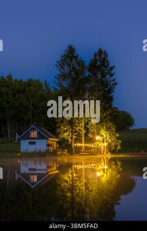 Eine gemütliche Hütte am See im ländlichen Estland, beleuchtet von warmen Außenlichtern, reflektiert in ruhigem Wasser in einer ruhigen Sommernacht. Stockfoto