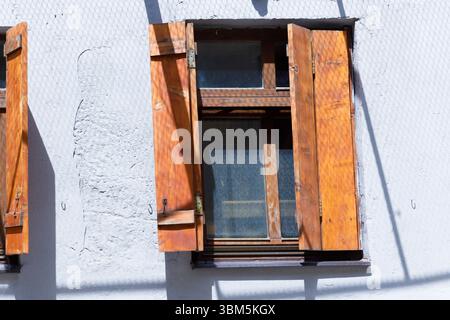 Dieses Bild zeigt ein elegant gefertigtes Fenster mit rustikalen Holzläden, das auf einer strukturierten weißen Wand positioniert ist. Das Zusammenspiel von Licht und Stockfoto