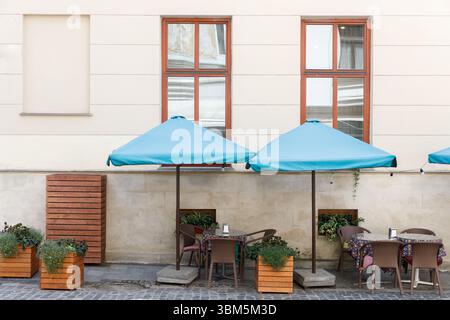 Das Bild zeigt ein malerisches Café im Freien mit blauen Sonnenschirmen, einladenden Tischen und üppigen grünen Pflanzgefäßen. Es ist ein perfekter Ort zum Entspannen und Entspannen Stockfoto