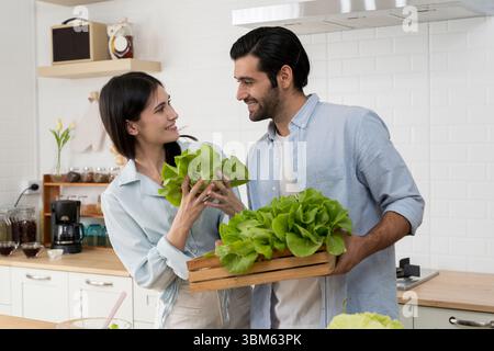 Glückliches Paar, das grünen Salat hält und in der Küche lächelt, während zusammen gesundes Essen zubereitet wird. Förderung von Wellness, Liebe, Teamwork und sauberem Essen Stockfoto