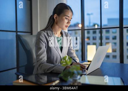 Ich arbeite allein spät in der Nacht. Asiatische Geschäftsfrau am Arbeitsplatz im Büro spät in der Nacht an einem Schreibtisch in einem dunklen Büro arbeiten an einem Laptop spät in der Nacht Stockfoto