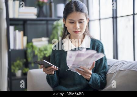 Asiatische Frau, die Papierdokumente hält, berechnet Geldeinsparungen und bezahlt Rechnungen in mobiler Anwendung mit Smartphone online mit zahlendem Haushalt Stockfoto