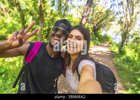 Interrassische Paare, die Rucksäcke tragen und ein Selfie machen, während sie durch einen üppigen Wald wandern und ihr Outdoor-Abenteuer genießen Stockfoto