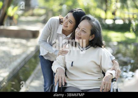 Asiatische Seniorin im Rollstuhl mit glücklicher Tochter. Familienbeziehung Rentnerin sitzend auf Rollstuhl in der Parkalpflege im Altersheim Stockfoto