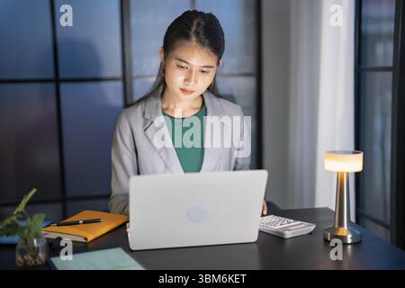 Ich arbeite allein spät in der Nacht. Asiatische Geschäftsfrau am Arbeitsplatz im Büro spät in der Nacht an einem Schreibtisch in einem dunklen Büro arbeiten an einem Laptop spät in der Nacht Stockfoto