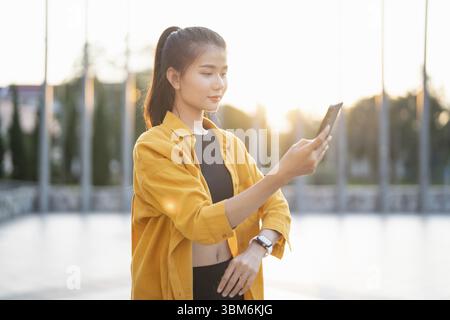 Lächelnde junge asiatische Frau, die ihr Smartphone in der Stadt benutzt und Apps im Freien überprüft Stockfoto