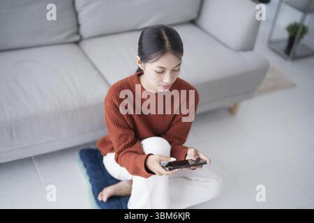 Asiatische Frau, die Smart Home kontrolliert. Mobiltelefon mit Smart-Home-App im Wohnzimmer. Touchscreen-Ai-Technologie. Fernbedienungssystem für Zuhause Stockfoto