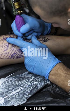 Vertikale Nahaufnahme der Hände eines Mannes, der in einem Tattoo-Studio das Bein einer Frau auf einer Bahre tätowiert Stockfoto