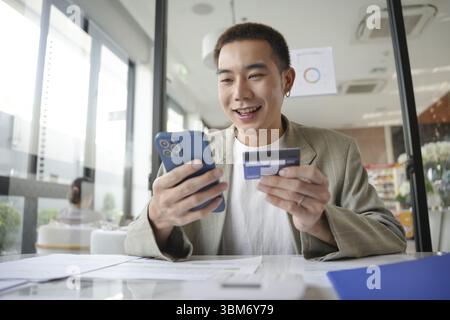 Asiatischer Mann, der mit Kreditkarte im Büro bezahlt. Mann, der Smartphone für Online-Shopping-elektronische Brieftasche benutzt Stockfoto