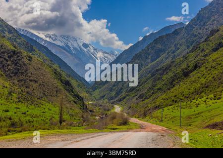 Atemberaubende Aussicht auf eine majestätische Bergkette unter einem sonnigen Himmel in Kirgisistan. Eine kurvenreiche Straße durchquert lebendige Vegetation und schafft ein ruhiges und ruhiges Ambiente Stockfoto