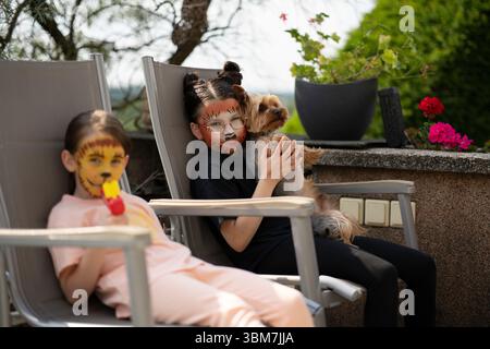 Zwei Schwestern mit tierischer Gesichtsfarbe sitzen auf Gartenstühlen in ihrem Garten. Konzept: Kinderfreude, Geschwisterbindung, Outdoor-Spaß, Fantasie, Familienmutter Stockfoto