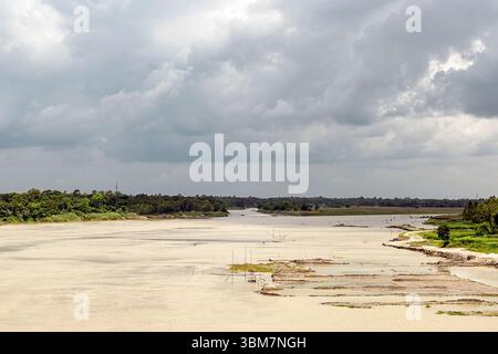 Ein sich windender Fluss schlängelt sich sanft durch eine ruhige ländliche Landschaft, flankiert von üppigen grünen Bäumen und offenen Feldern. Der bewölkte Himmel, gefüllt mit grauer Wolke Stockfoto