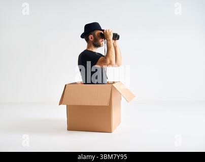Mann mit Hut, der durch das Fernglas schaut, sitzt in einem Karton. Spion, lustiges Konzept Stockfoto
