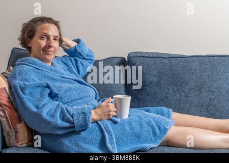 Eine Frau in einem blauen Gewand sitzt auf einer Couch mit einer Tasse in der Hand. Sie ist entspannt und bequem Stockfoto
