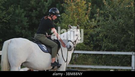 Junge Reiterin mit Helm und lässiger Kleidung, die auf ihrem weißen Pferd sitzt und es nach einem Training im Reitstall streichelt Stockfoto