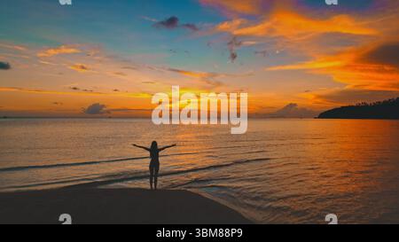 Silhouette einer jungen Frau, die mit offenen Armen an einem Strand steht und die lebendigen Farben eines atemberaubenden Sonnenuntergangs über dem ruhigen Meer in Thailand, Asien, genießt Stockfoto