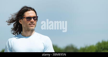 Glücklicher, selbstbewusster Mann, brünett mit langen lockigen Haaren, die im Wind in einer Sonnenbrille wehen und wegschauen. Banner. Sommer. Naturhintergrund für Reflexion. Stockfoto