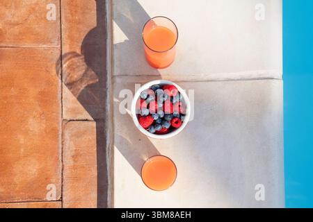 Blick von oben nach unten auf ein gesundes Frühstück mit einer Schüssel frischer Beeren und Gläsern Saft am Rand eines Swimmingpools an einem sonnigen Tag Stockfoto