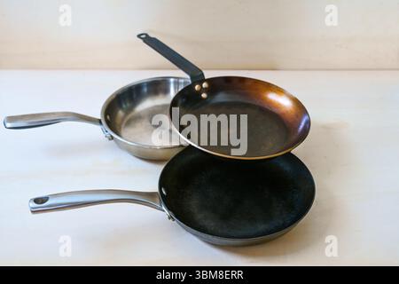 Drei Pfannen ohne ungesunde Antihaftbeschichtung aus Kohlenstoffstahl, Edelstahl und Gusseisen auf hellem Holzhintergrund, Kopierraum, se Stockfoto